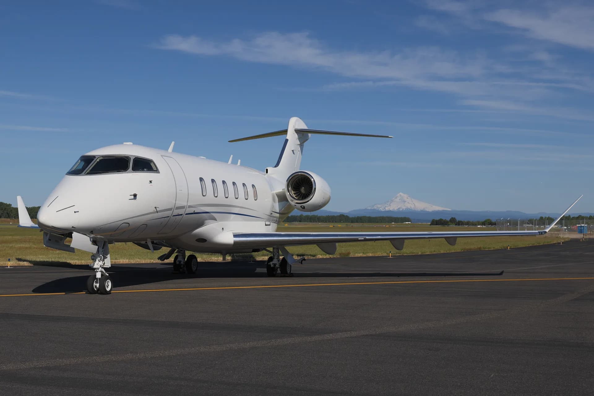A white Bombardier Challenger business jet parked on an airport tarmac, with a snow-capped mountain in the background.