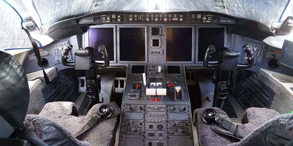 The cockpit of a private jet features a complex array of digital displays, control panels, and yokes, with sheepskin covers on the pilot seats.