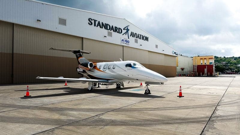A Phenom 100 aircraft is parked in front of a Standard Aviation hangar.