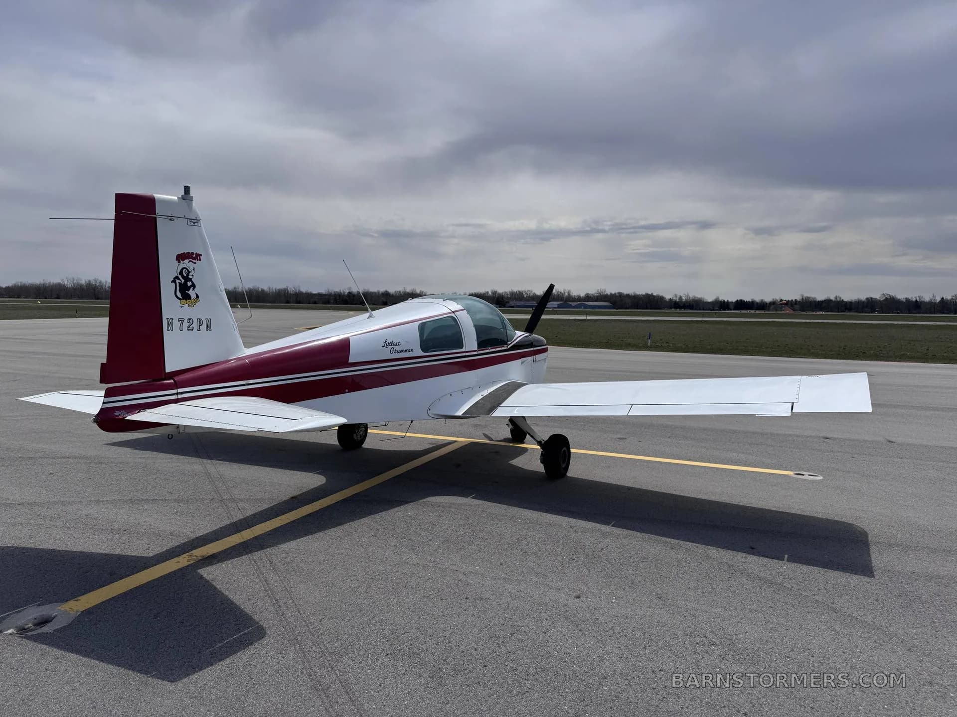 A white and maroon Grumman American AA-5 Traveler aircraft is parked on an asphalt tarmac under a cloudy sky.