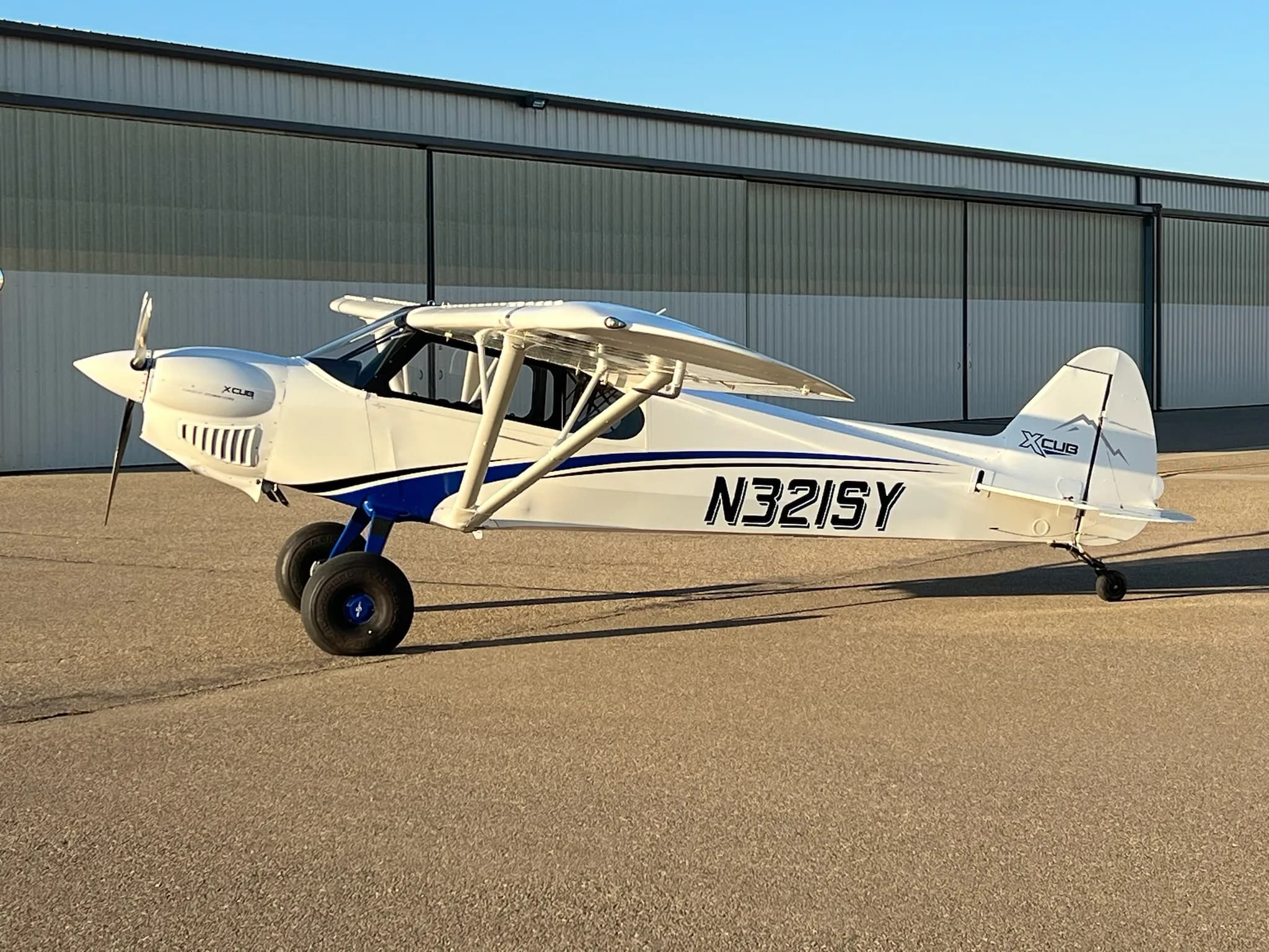 A white X-Cub aircraft with blue stripes and registration N321SY parked on an airfield in front of a hangar.