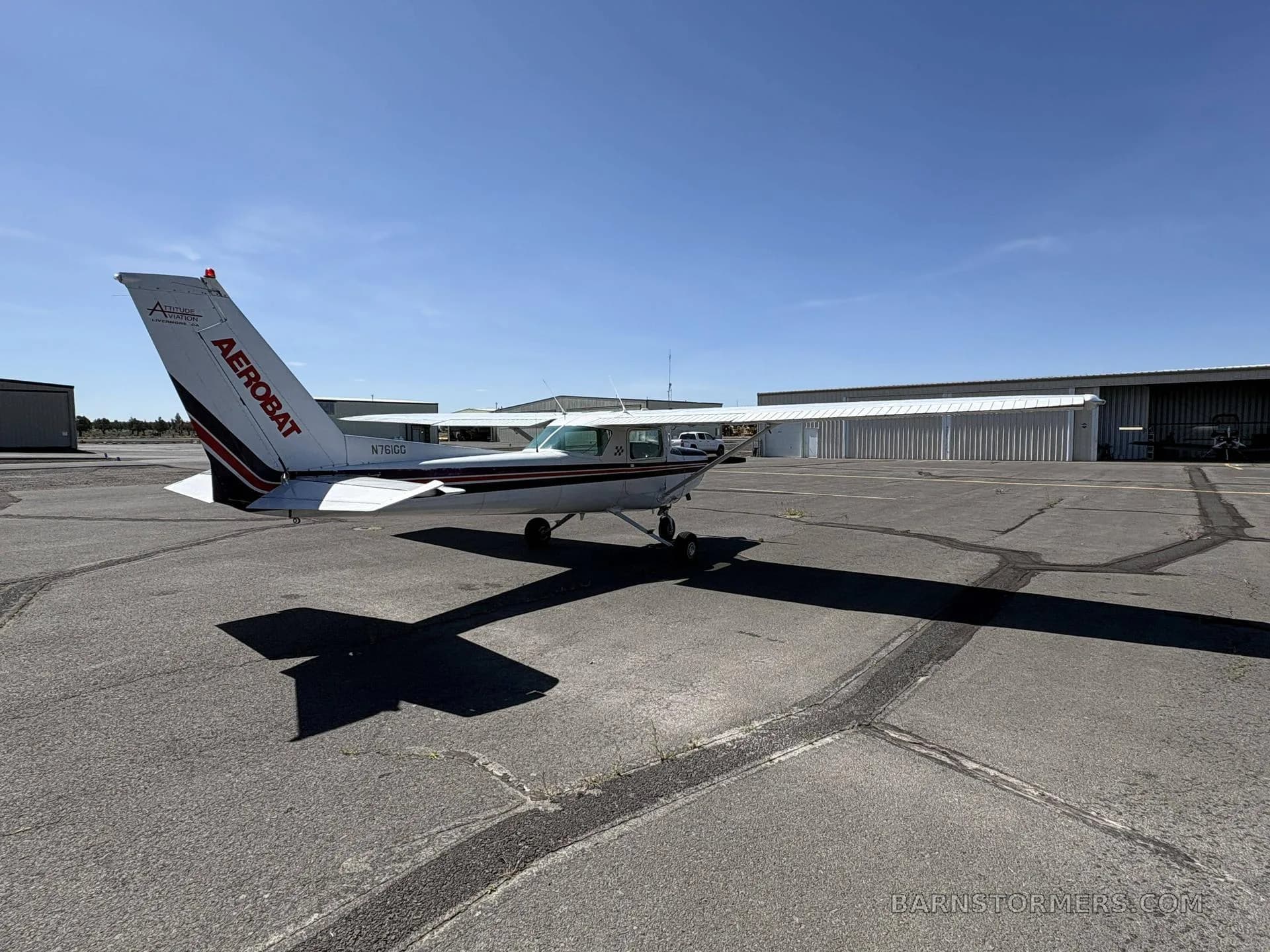 A rear-quarter view of a white and maroon Cessna 150 Aerobat (N761GG) parked on an asphalt apron under a clear sky.