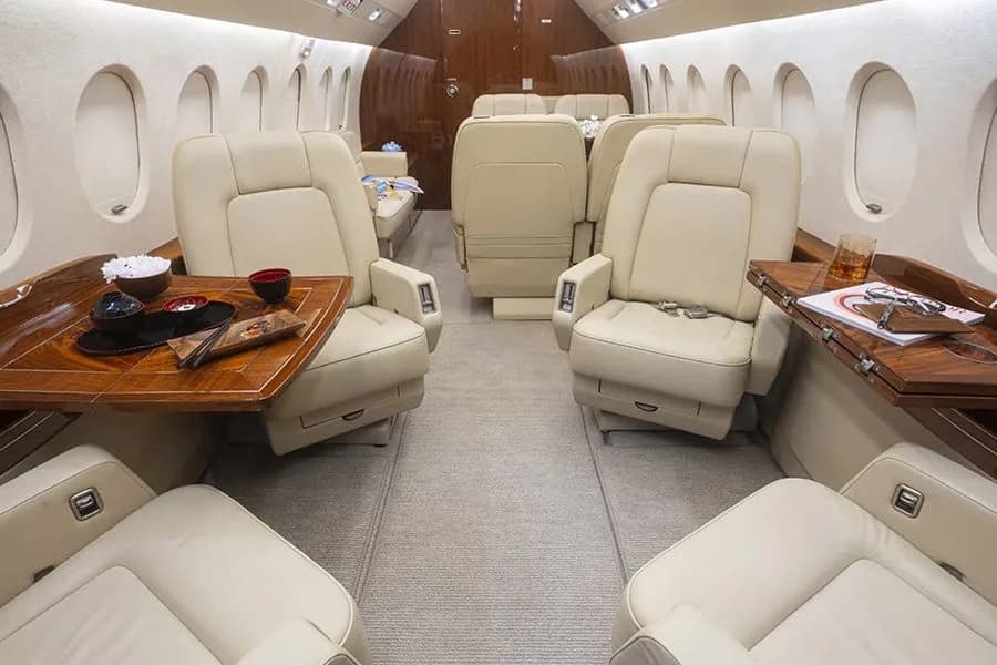 The spacious interior of a private jet cabin, featuring multiple cream-colored leather seats and two polished wooden tables with items on them.