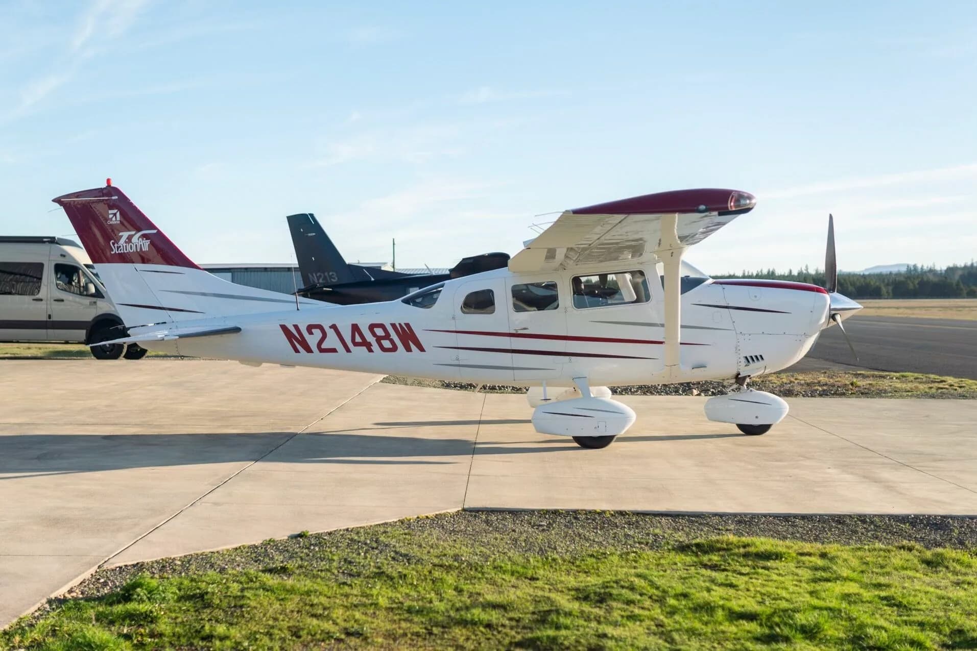 A white Cessna aircraft with red and dark red stripes, registration N2148W, parked on a tarmac with other vehicles and aircraft in the background.