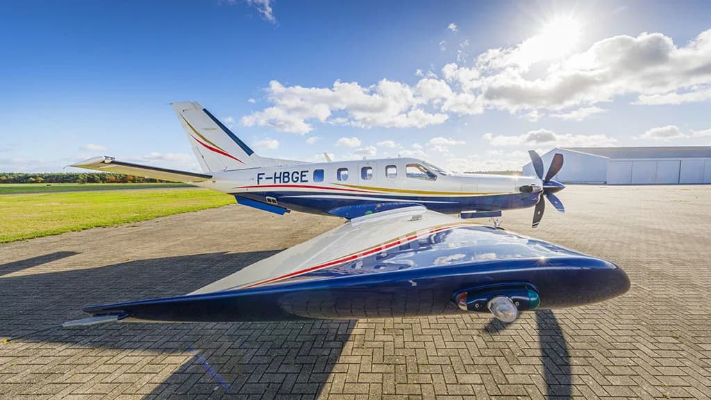 A white and blue TBM aircraft with registration F-HBGE parked on a paved surface, with the sun shining brightly in the background.