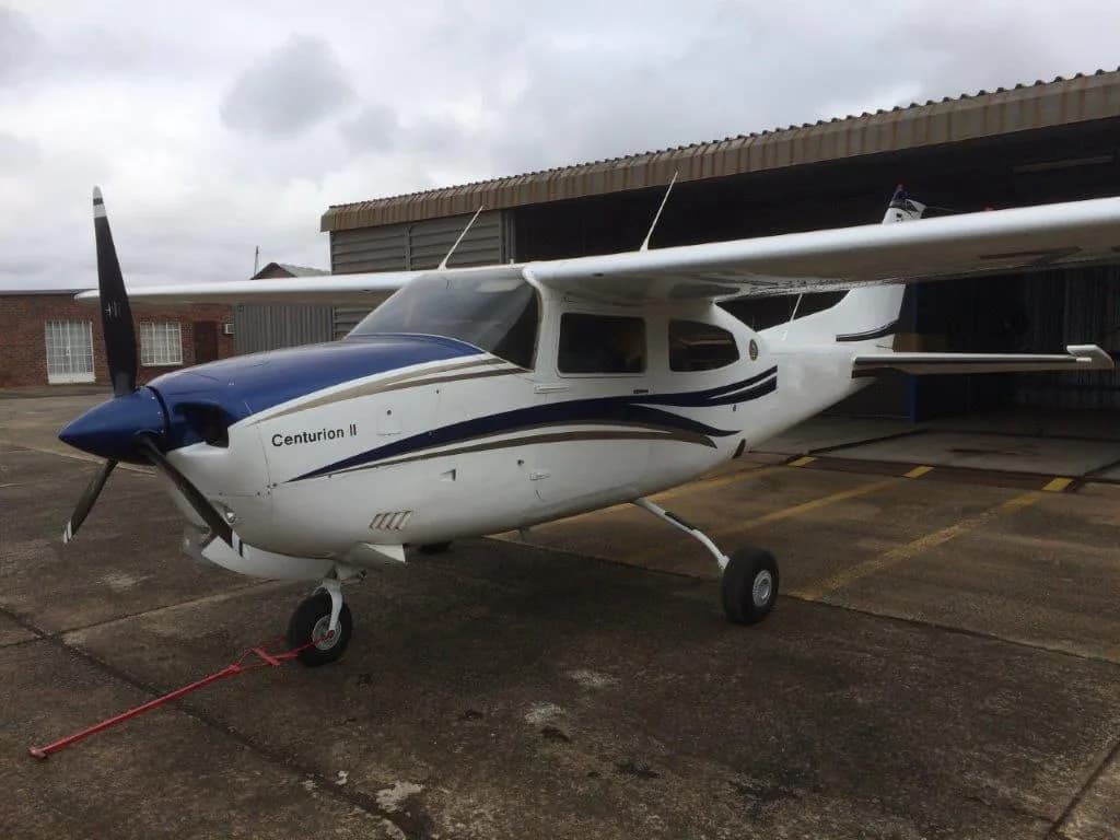 A white Cessna Centurion II with blue and gold stripes is parked on an asphalt apron next to a hangar under a cloudy sky.