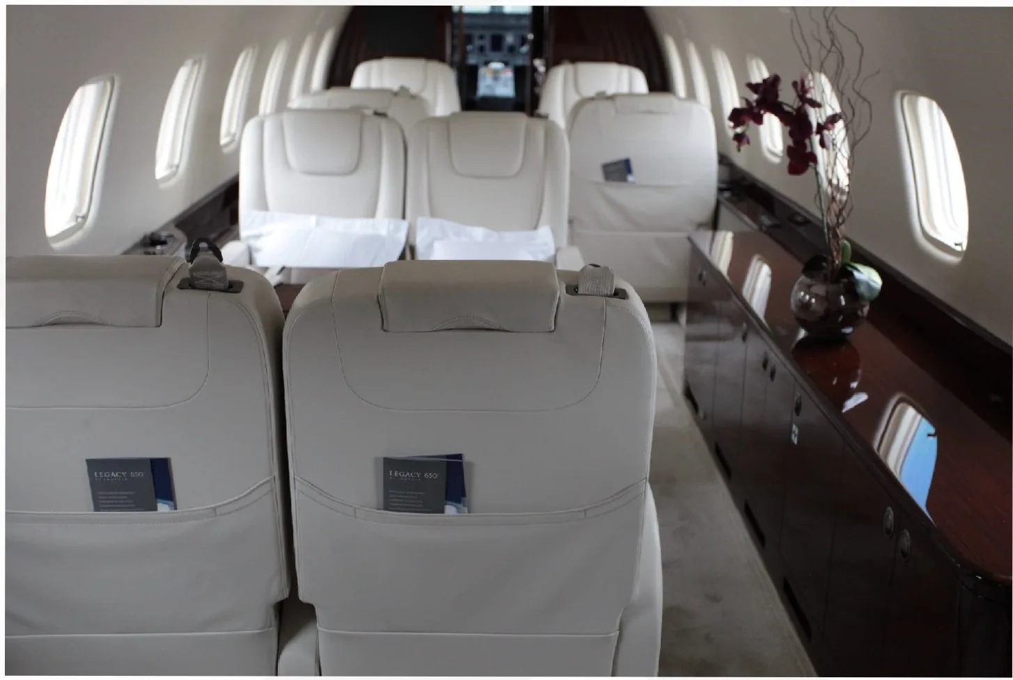 The interior of a private jet cabin with cream-colored leather seats, dark wood cabinetry, and a decorative vase with flowers.