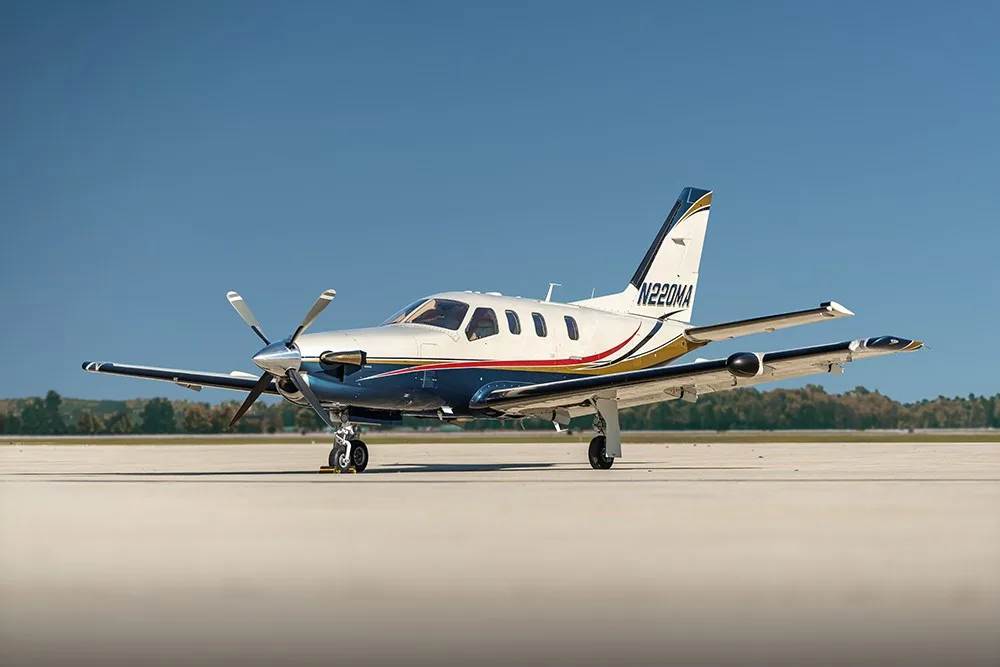 A white, blue, and gold painted turboprop aircraft (N220MA) parked on an airfield, viewed from a slightly lower angle.
