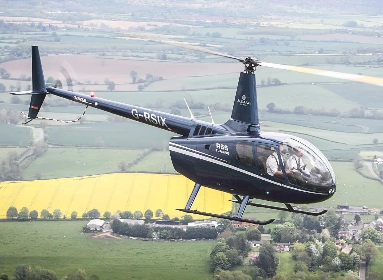A blue Robinson R66 Turbine helicopter in flight over a patchwork of fields.