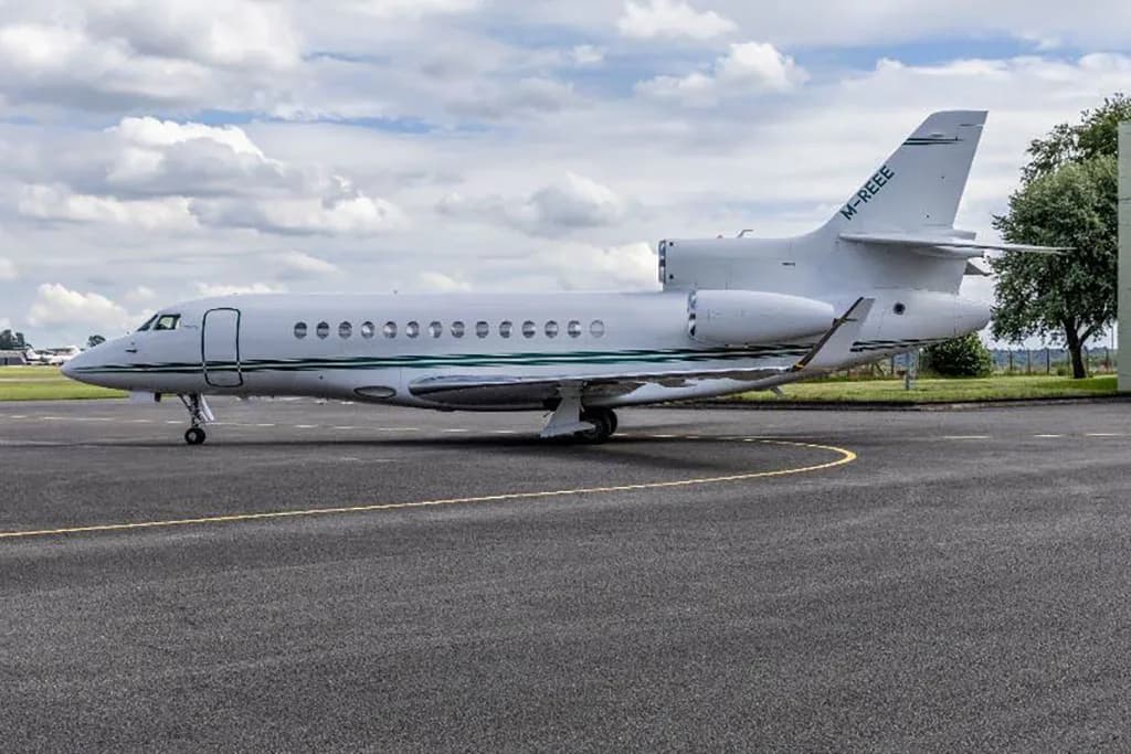A side view of a white Dassault Falcon 900EX with a green stripe, registration M-REEE, parked on an airfield.