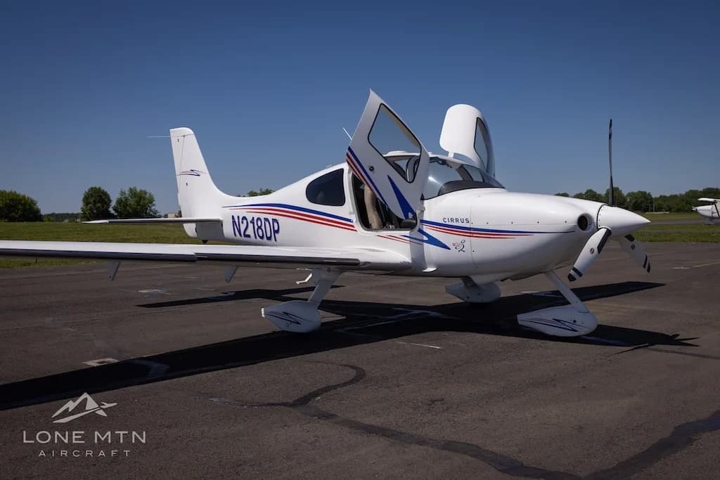 Front-right view of a white Cirrus aircraft (N218DP) with blue and red stripes, doors open, parked on an airfield.