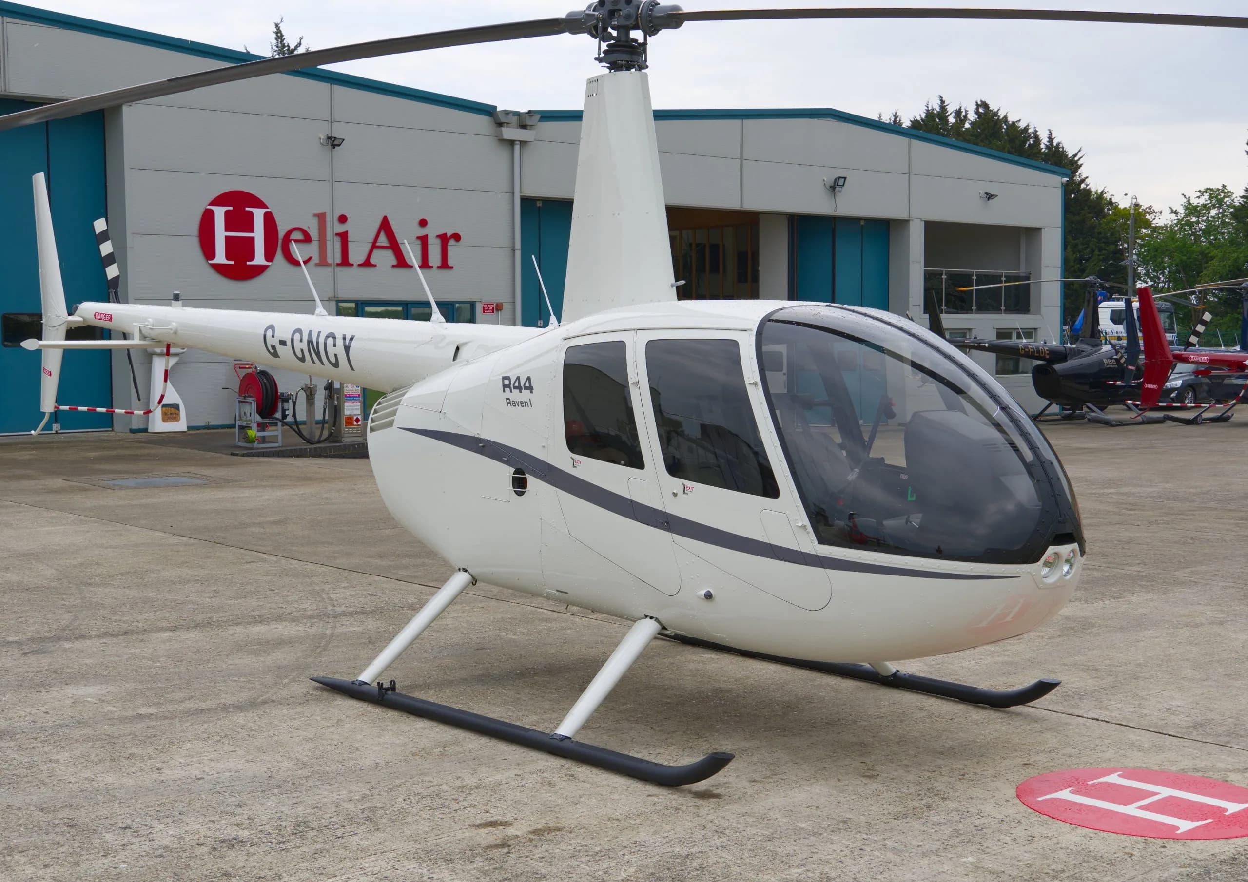 A white Robinson R44 Raven I helicopter (G-GNGY) with dark grey stripes parked on a tarmac in front of a Heli Air hangar.