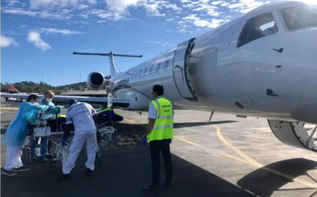 A white regional jet is on the tarmac with medical personnel loading a stretcher into its open door under a blue sky.