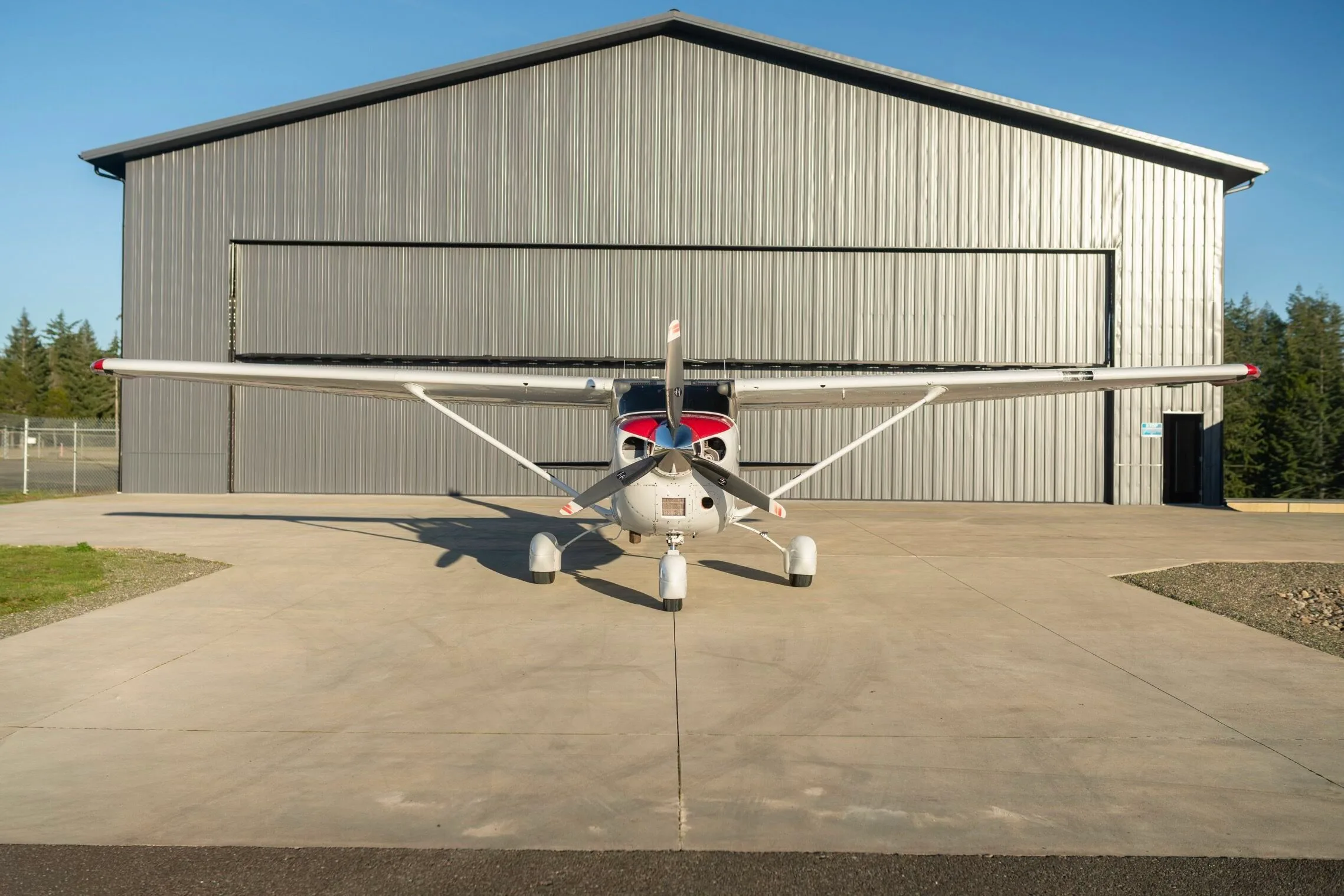 A white Cessna aircraft with red accents on the nose, registration N2148W, seen from the front, parked in front of a large metal hangar.