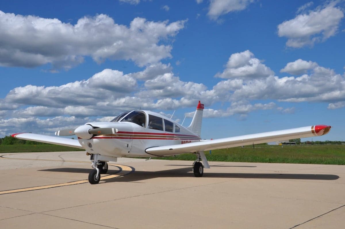 A white single-engine aircraft with red and gold stripes parked on an airfield with a blue sky and clouds.