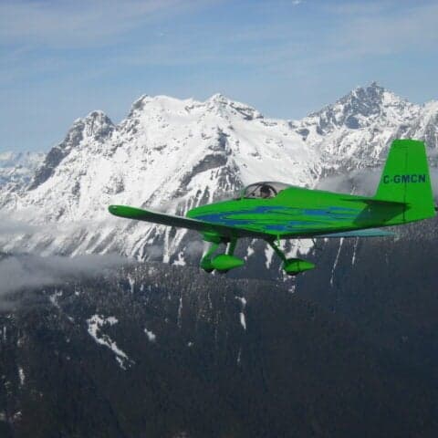 A green aircraft with blue accents flying in front of snow-covered mountains.