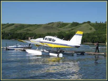 A white and yellow single-engine floatplane is docked on a lake with green hills in the background.