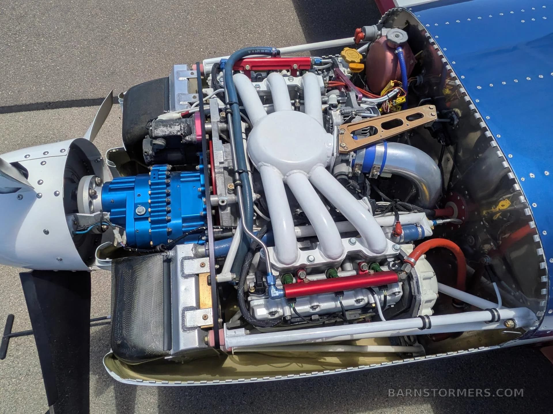 The exposed engine bay of the aircraft reveals a complex engine with white exhaust headers, a blue alternator, and various hoses and components.