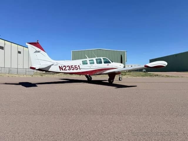 A white and red Beechcraft Bonanza aircraft is parked on an asphalt runway with a hangar in the background.