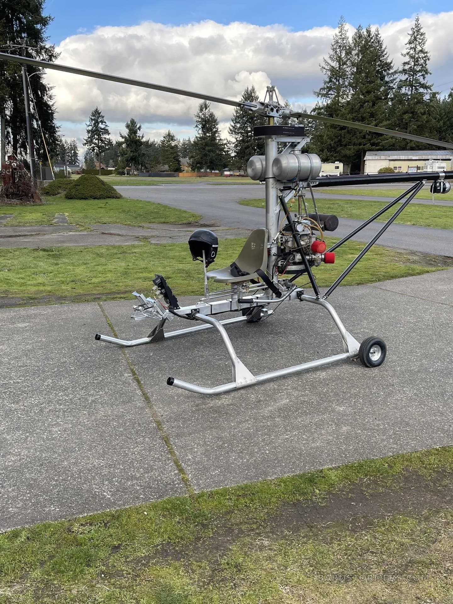 A single-seat ultralight helicopter with an open frame and a black helmet resting on the seat, parked on concrete with trees in the background.