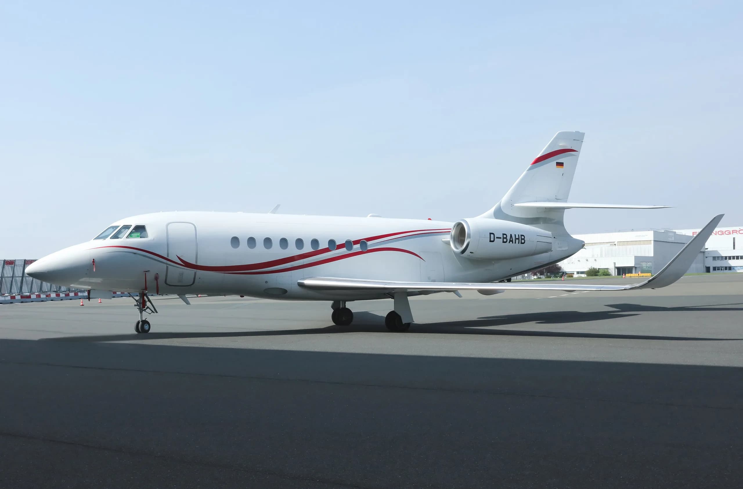A white business jet with red and grey stripes and German flag on the tail, parked on an airport apron.