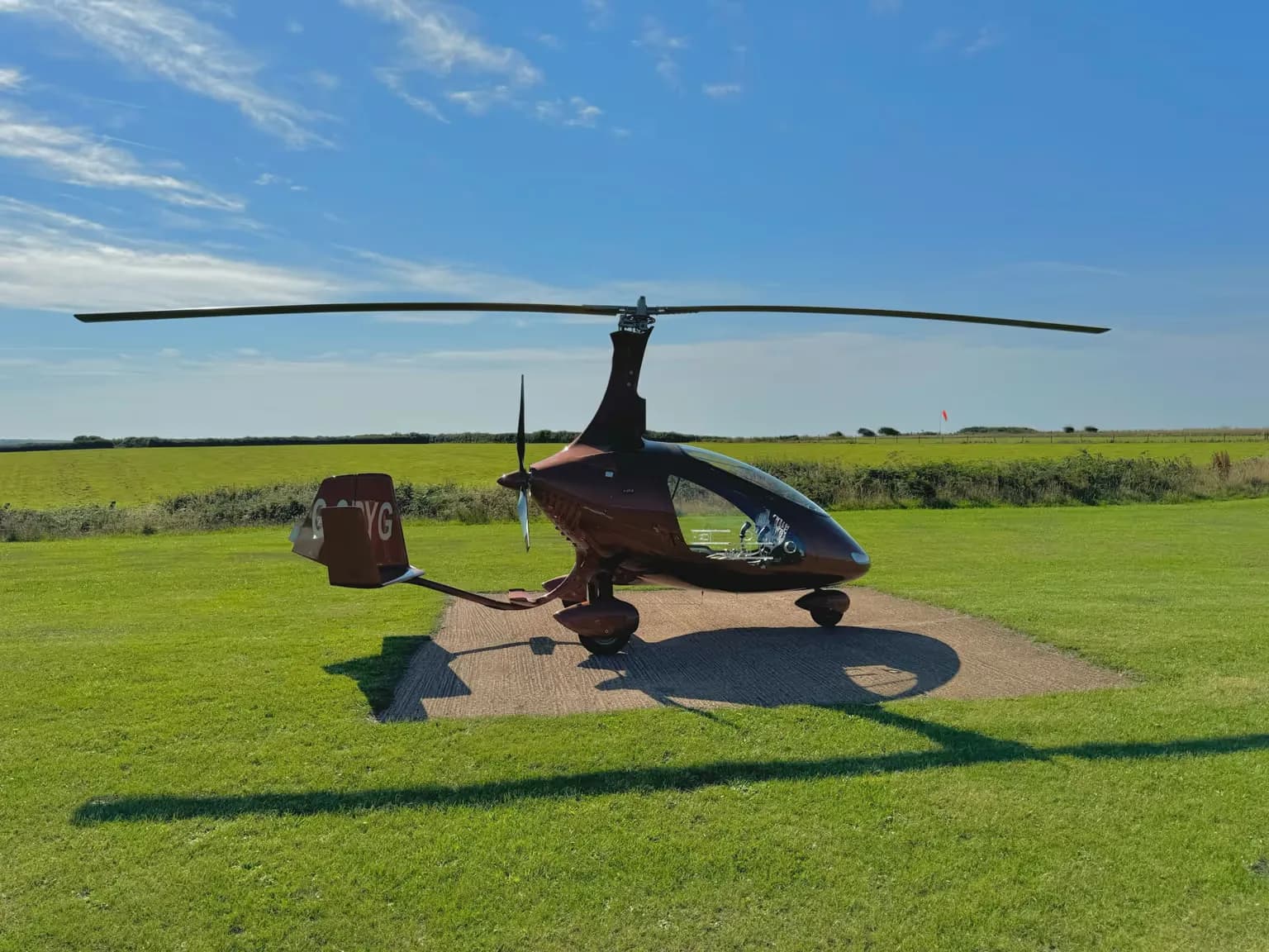 A brown gyrocopter, G-ORYG, parked on a concrete pad in a grassy field under a blue sky.