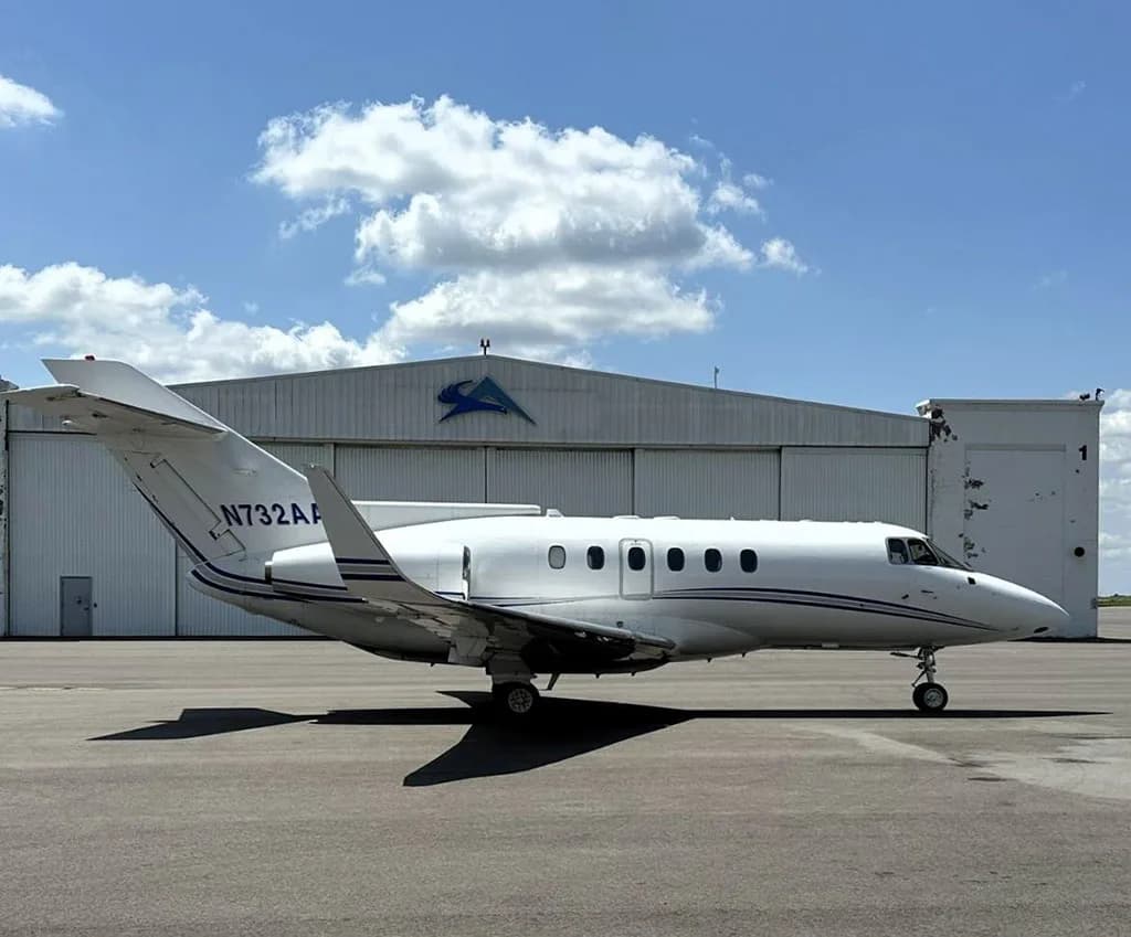 A white business jet, N732AA, with blue stripes parked on an tarmac in front of a hangar under a partly cloudy sky.