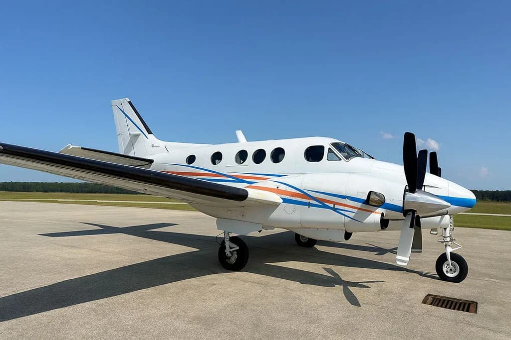A white twin-engine turboprop aircraft with blue and orange stripes, parked on an airfield with a clear blue sky.