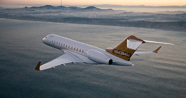 A white Bombardier Global business jet with gold and brown accents flying over a coastline.