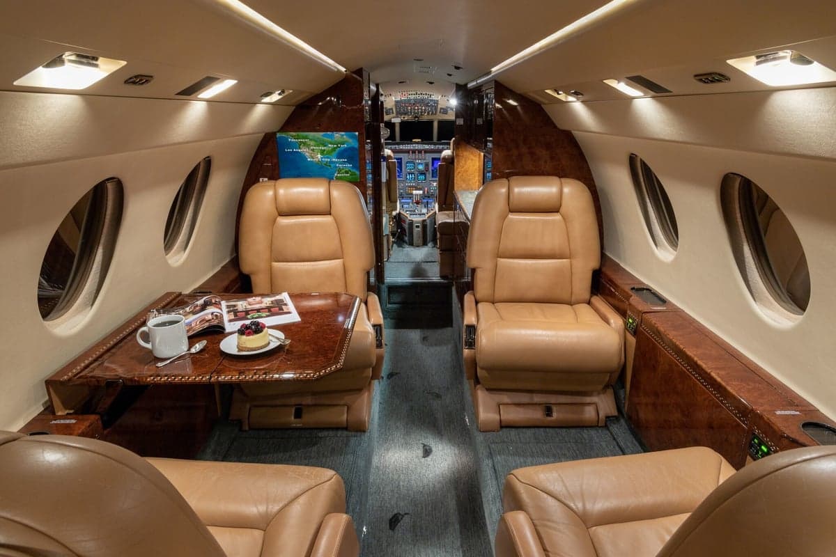 The luxurious interior cabin of a private jet featuring tan leather seats, a wooden table, and a map displayed on a screen.