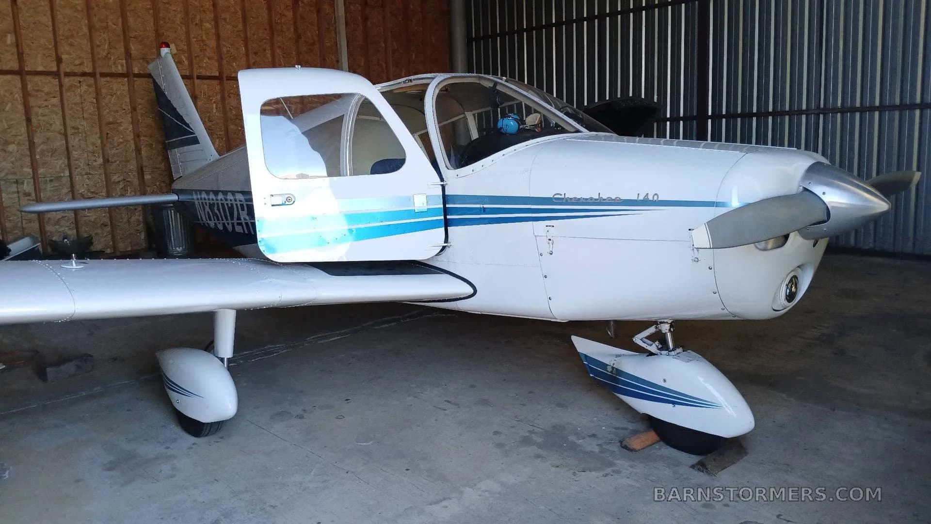 A white and blue Piper Cherokee 140 (N8302R) with open doors, parked inside a hangar.