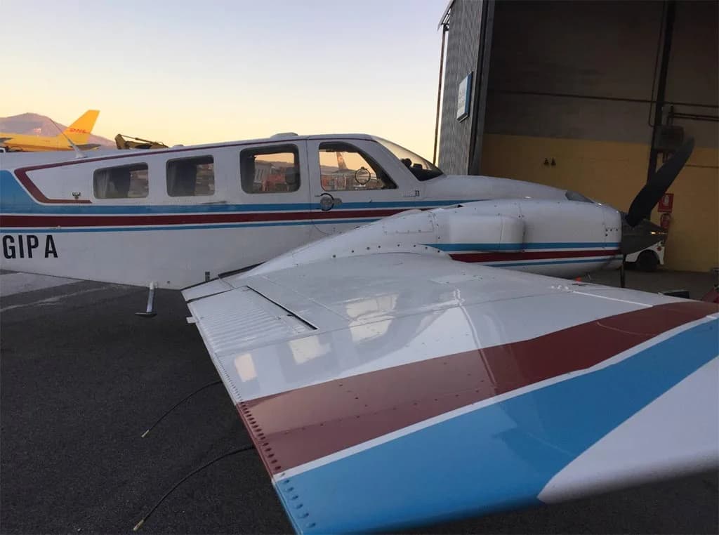 Side view of a white twin-engine aircraft with blue and red stripes, registration I-GIPA, parked next to a hangar at sunset.