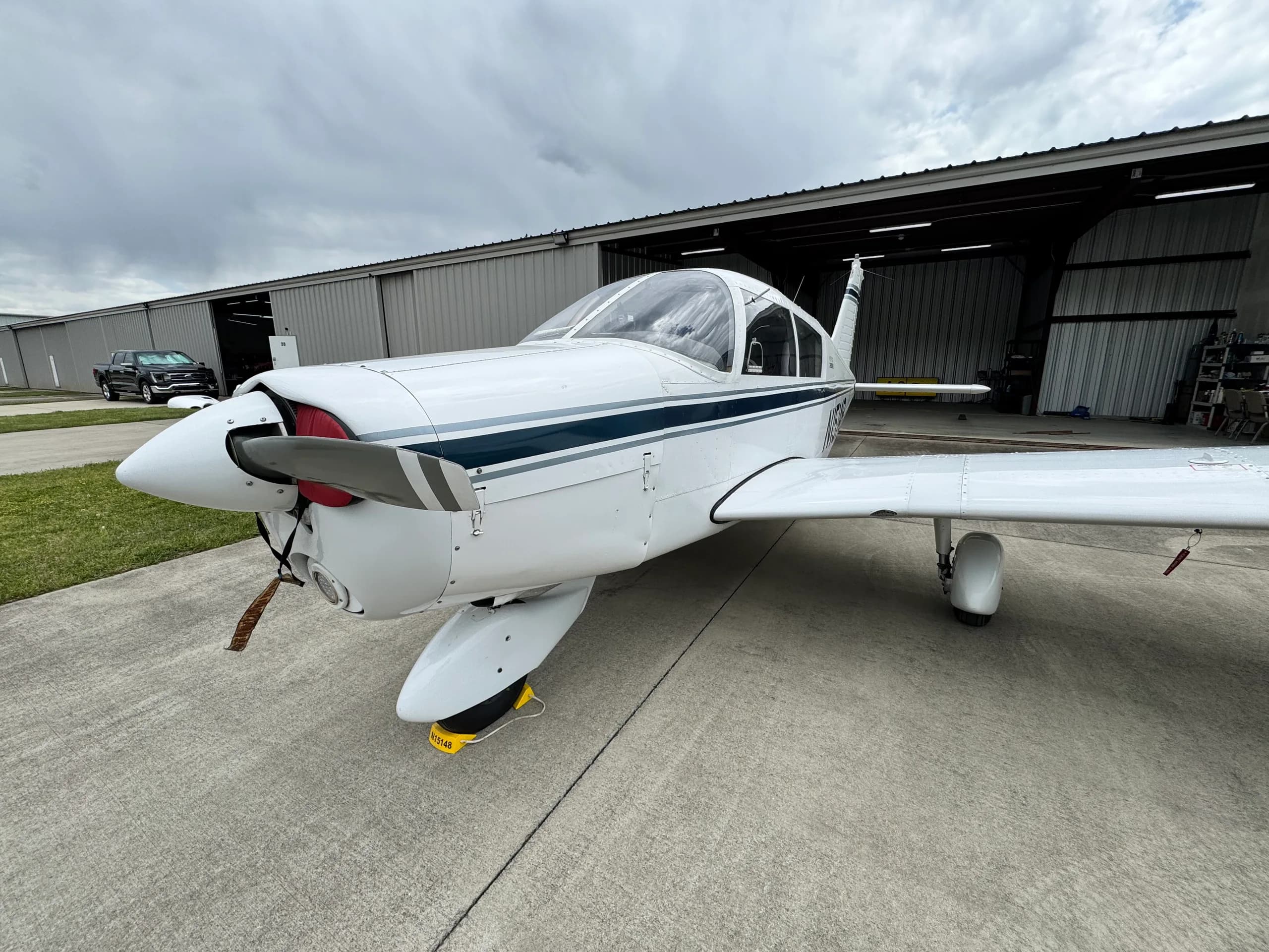 Front-quarter view of a white single-engine aircraft, N15148, with dark blue stripes, parked on concrete in front of a hangar.