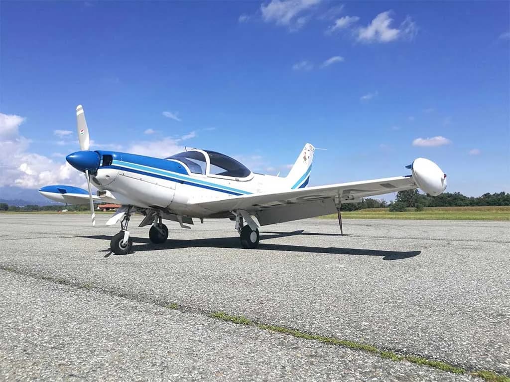 A white and blue single-engine aircraft with wingtip tanks is parked on an asphalt runway under a blue sky.