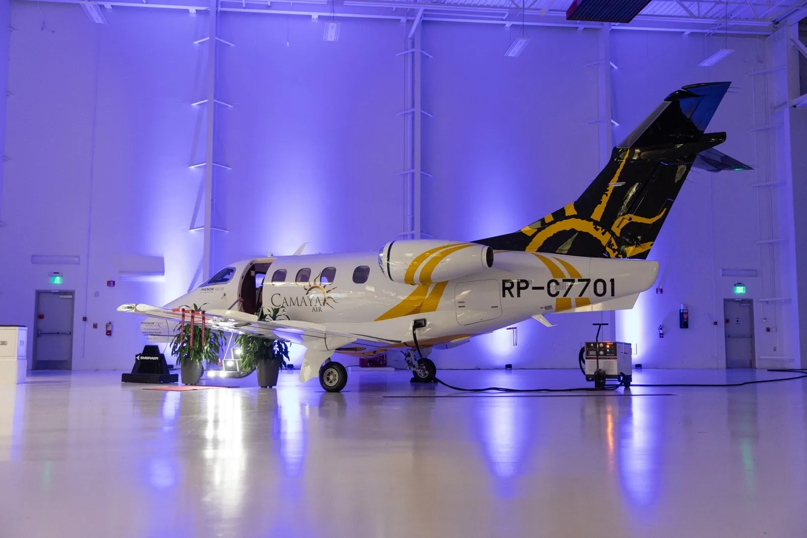 The Camaya Air Embraer Phenom 300, registration RP-C7701, displayed in a hangar with blue lighting.