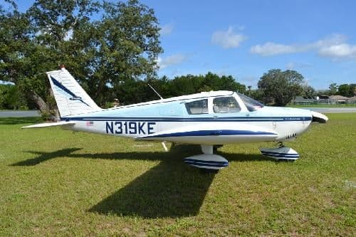 A white and blue Piper PA-28-140 Cherokee is parked on the grass.