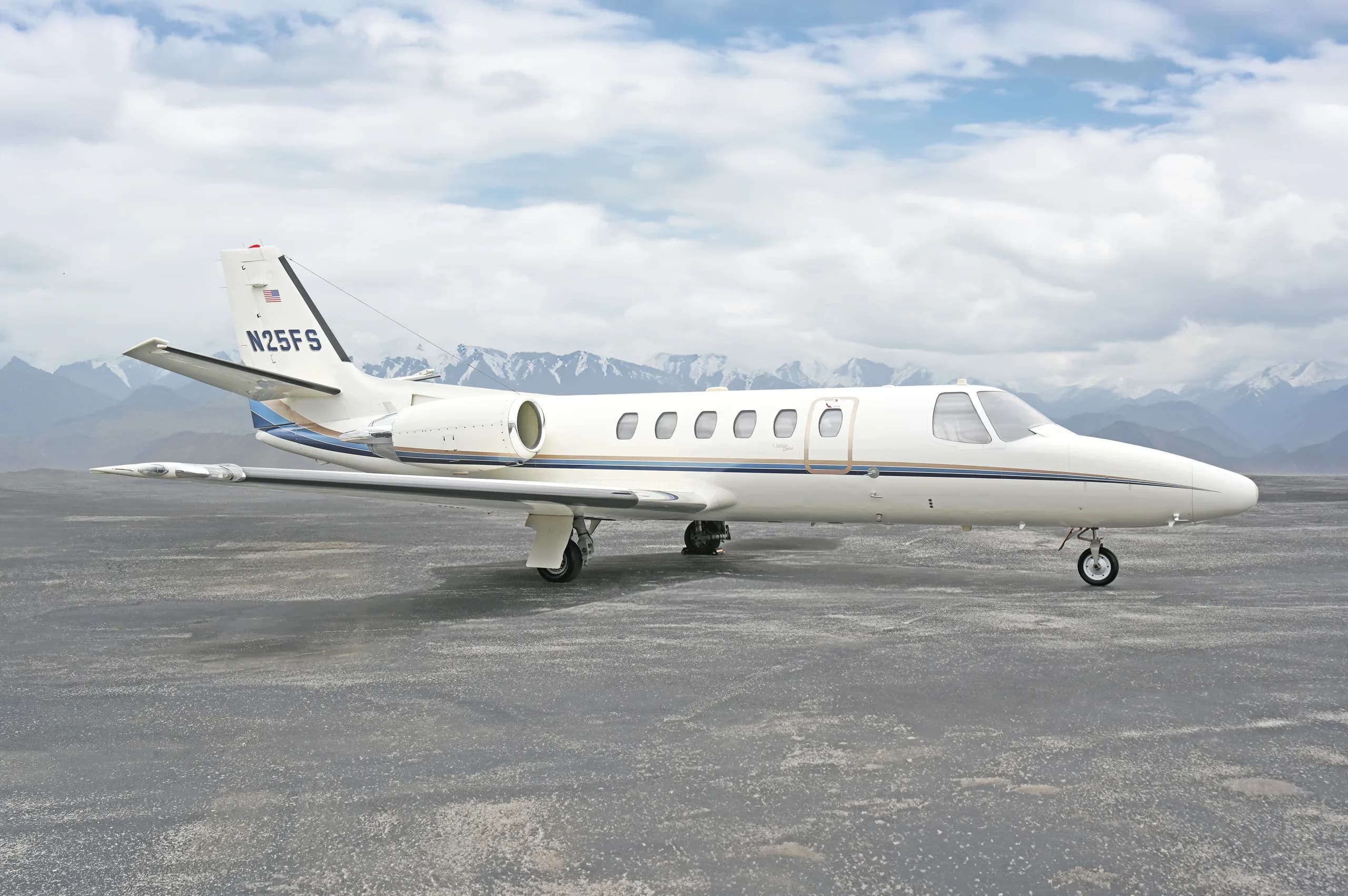 A white Cessna Citation business jet (N25FS) with blue and gold stripes, parked on an asphalt surface with snow-capped mountains in the background.