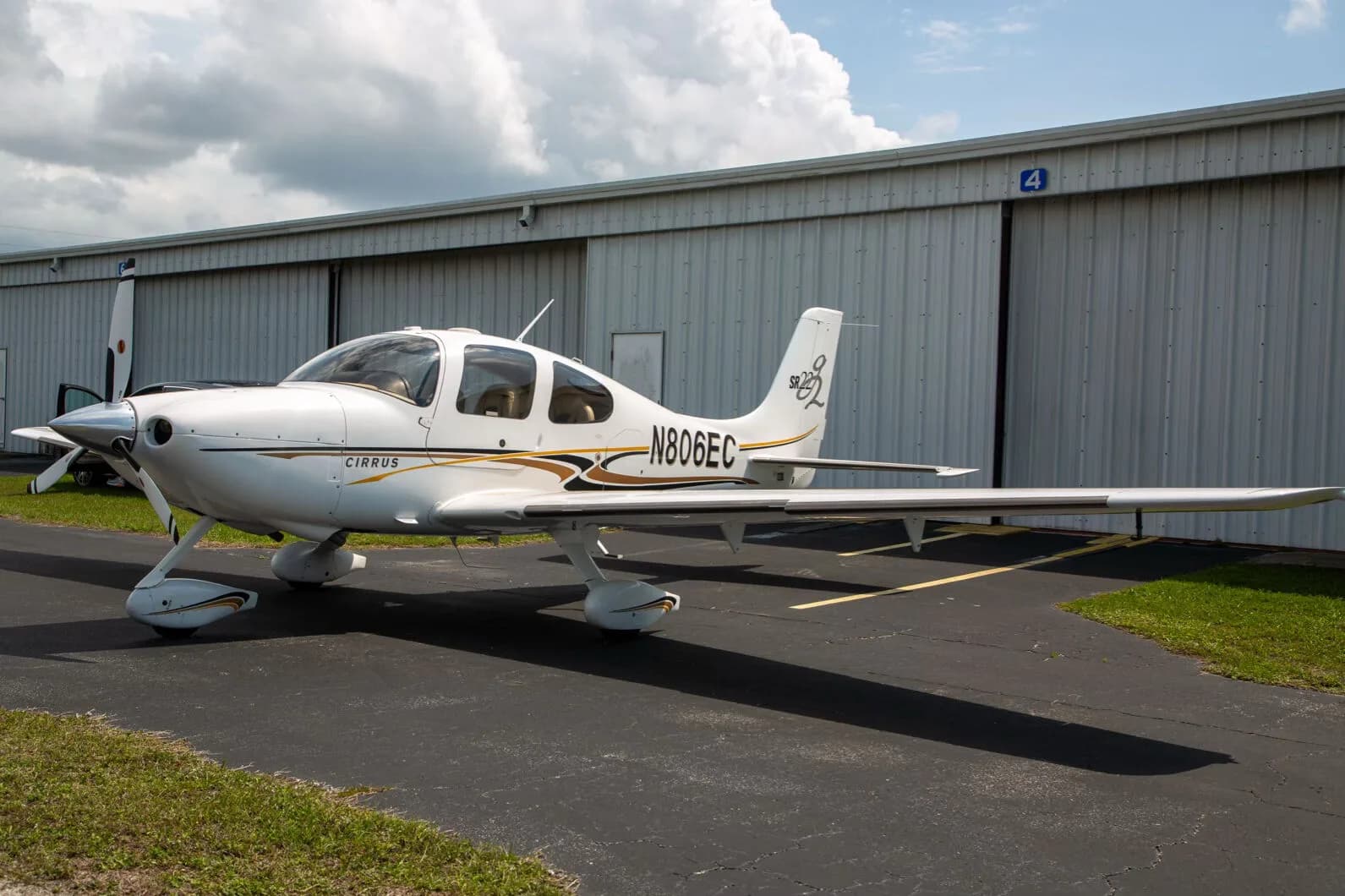 A white Cirrus SR22 aircraft with black and gold stripes, registration N806EC, parked on an asphalt tarmac in front of a hangar.