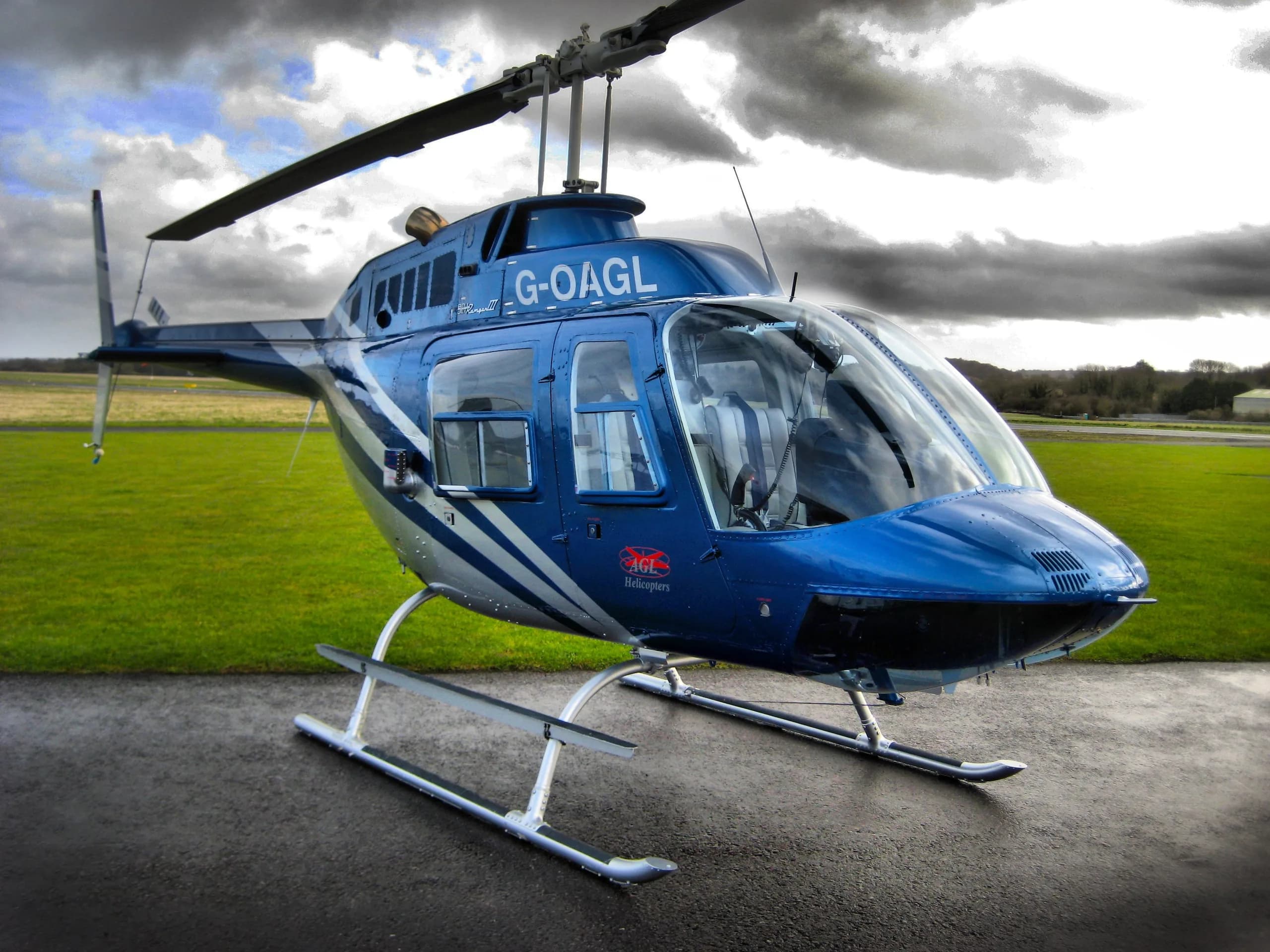A blue and silver Bell 206 JetRanger helicopter (G-OAGL) sits on a wet tarmac next to a green field under a cloudy sky.