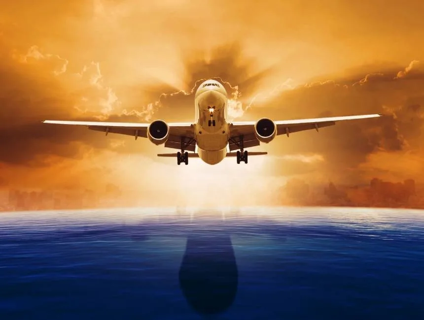 A commercial airliner is shown head-on, taking off over water at sunset.
