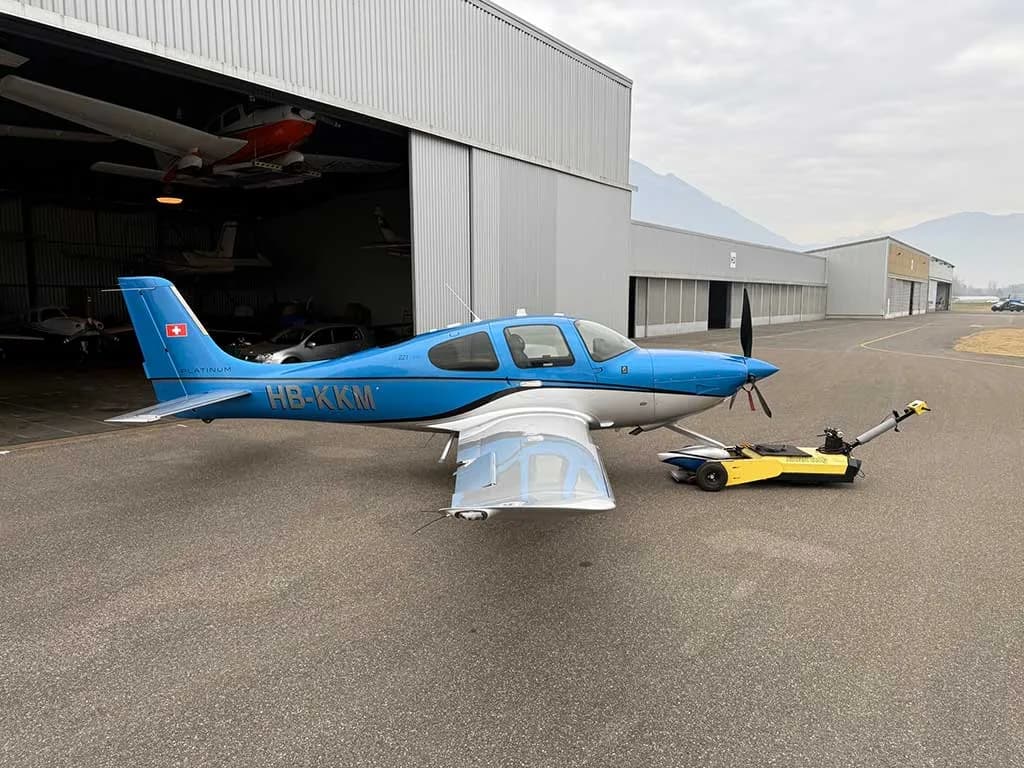 A side view of a blue and silver Cirrus SR22T aircraft with registration HB-KKM parked outside a hangar.