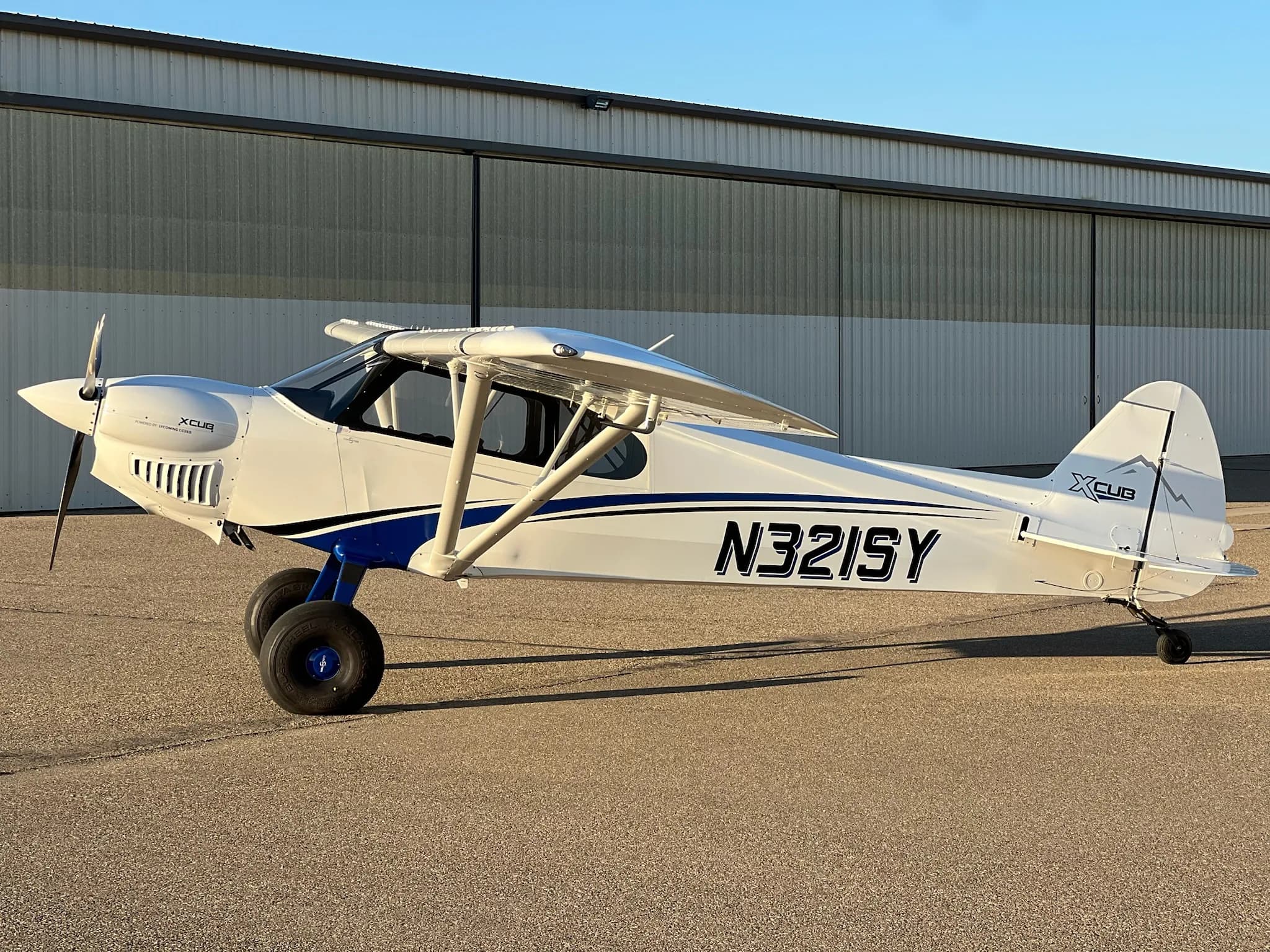 A white X-Cub aircraft with blue stripes and registration N321SY parked on an airfield in front of a hangar.