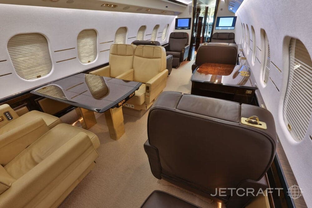 The spacious business jet cabin with a mix of brown and beige leather seating arrangements and a large dark table.