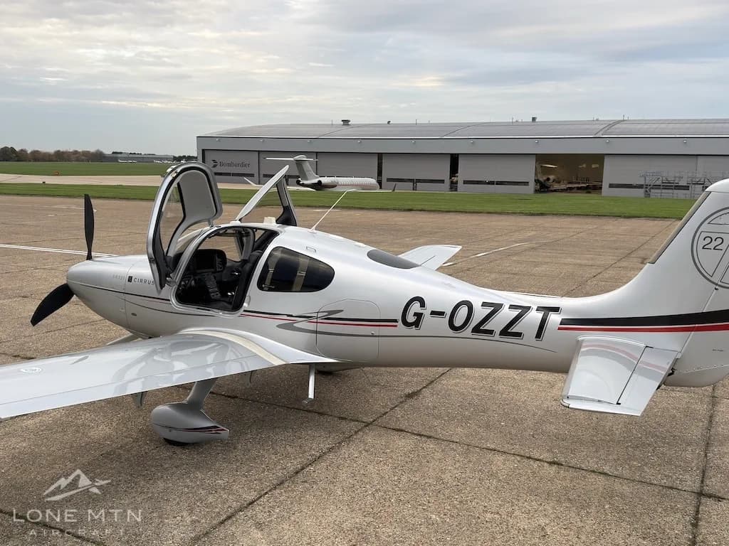 A silver Cirrus SR22T aircraft with registration G-OZZT and open door, parked on an airfield tarmac.
