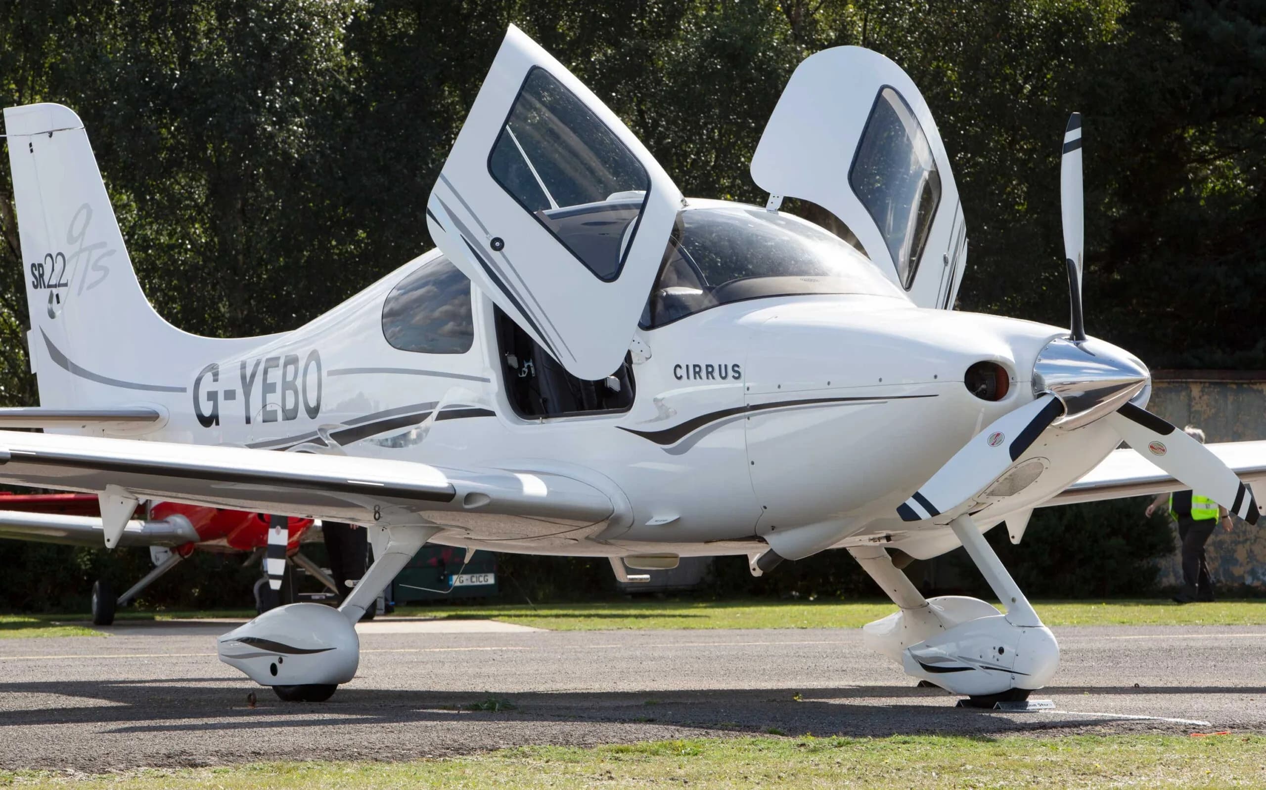 A white Cirrus SR22 TSi aircraft with black and grey stripes, registration G-YEBO, parked on an airfield with its gull-wing doors open.