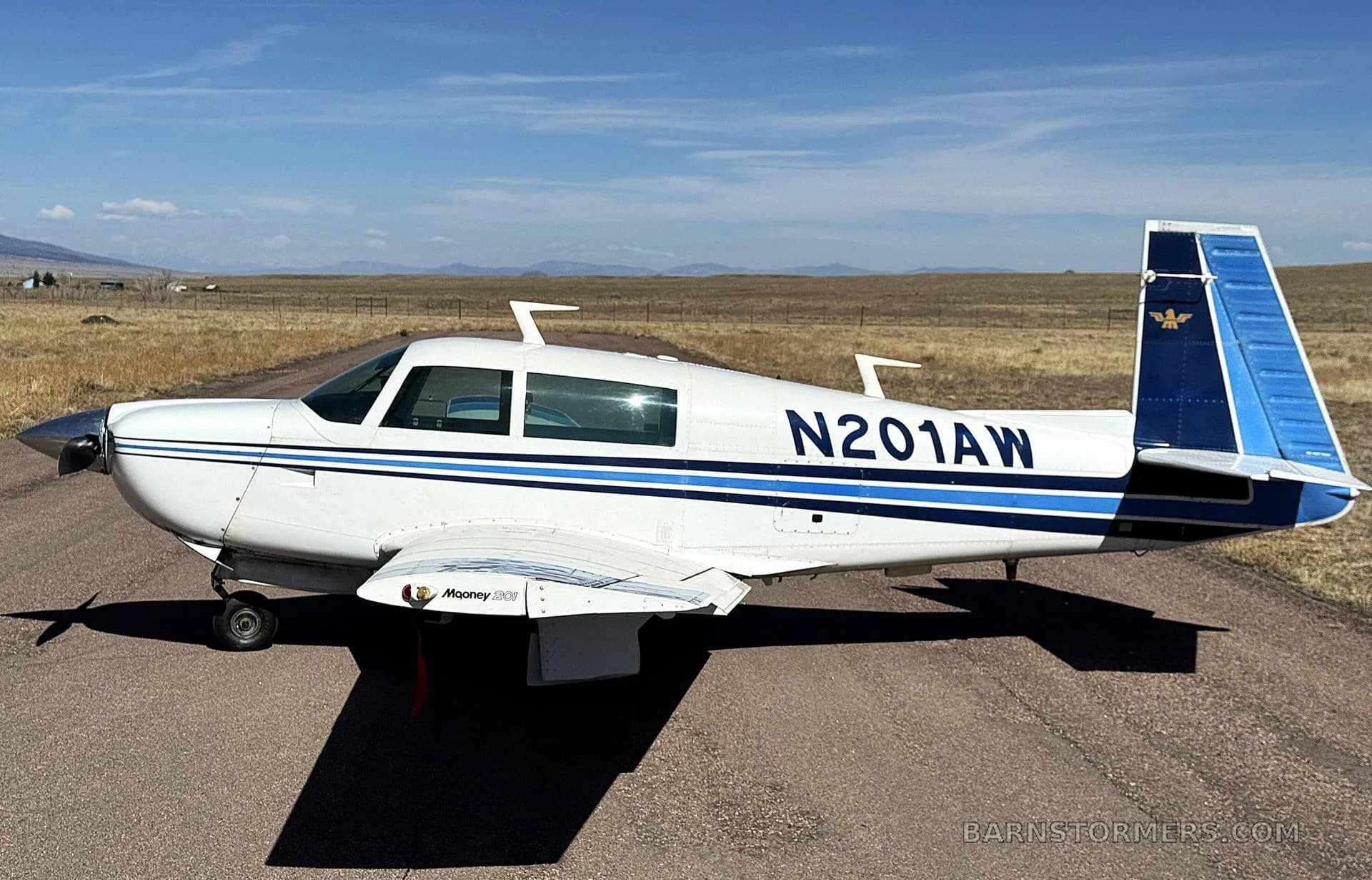 A white Mooney M20J aircraft with blue stripes and tail, registration N201AW, parked on an asphalt runway with mountains in the background.