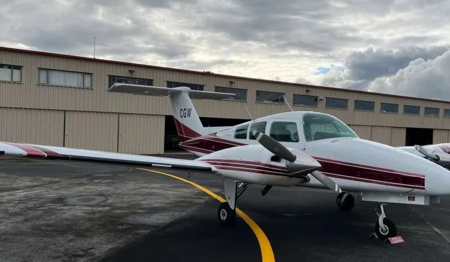 A white twin-engine propeller aircraft with maroon stripes is parked on an tarmac in front of a hangar under a cloudy sky.