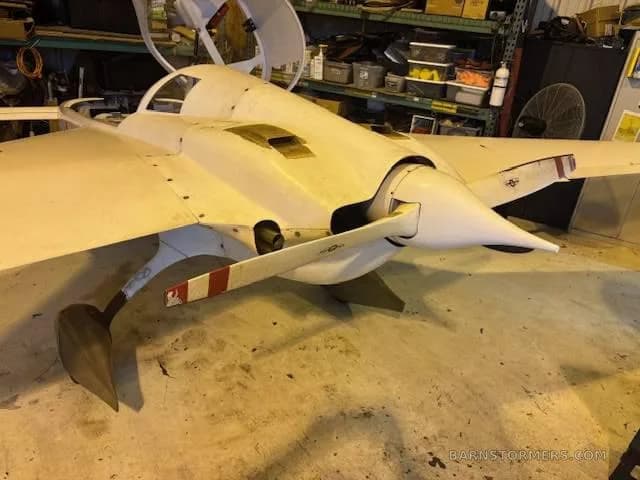 A white Rutan Long-EZ experimental aircraft with a pusher propeller parked inside a cluttered hangar.