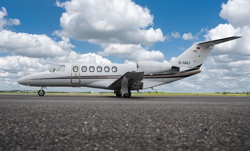 A white Cessna Citation business jet with black and brown stripes, registration D-IALI, parked on an asphalt runway under a blue sky with clouds.