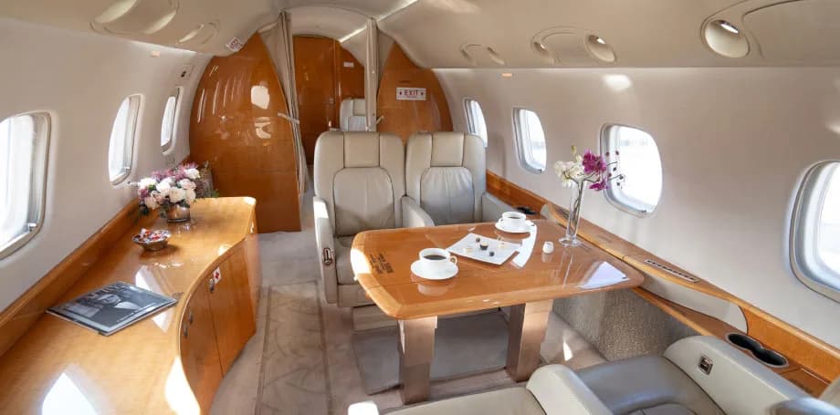 A private jet cabin interior featuring a dining table set with coffee cups and flowers, surrounded by light leather seats.
